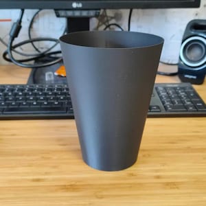 Op de afbeelding: Een zwarte, taps toelopende cilindrische container. De container is gemaakt van een mat materiaal en staat op een houten bureau. Een computertoetsenbord, muis en luidsprekers staan op de achtergrond.