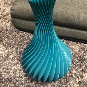 Op de afbeelding: Een turquoise vaas met een uniek, spiraalvormig ontwerp. De vaas heeft een brede basis die taps toeloopt naar een smalle hals, met een gerimpelde bovenrand. Het spiraalpatroon creëert een textuur, wat visuele interesse toevoegt.