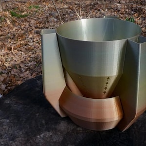Op de afbeelding: Een decoratieve vaas met een cilindrisch interieur en een hoekige buitenkant. De vaas heeft een bronzen basis die overgaat in een goudgroene top. Het ontwerp bevat een gebogen, bronzen accent en pijldetails. De vaas is gemaakt van een hard materiaal.
