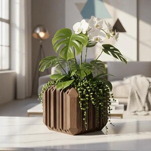 Op de afbeelding: Een donkerbruine houten plantenbak met een geometrische, verticaal geribbelde buitenkant. De bak bevat een weelderig arrangement van groene monstera-bladeren, een hangende erwtenplant en een tak met witte orchideeën.