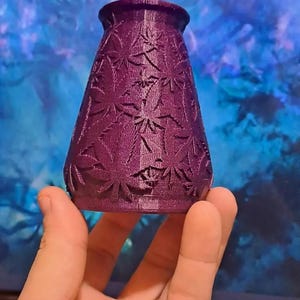 Op de afbeelding: Een paarse vaas met een uitlopende bovenkant en uitsparingen van cannabisbladeren. De vaas wordt in een hand gehouden. De achtergrond is een wazig blauw en wit.
