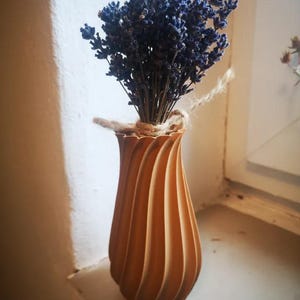 May include: A wooden vase with a spiral design holds a bundle of dried lavender. The vase is a warm brown colour and sits on a windowsill. The lavender has dark purple flowers and is tied with twine.