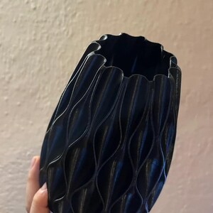May include: A black vase with a repeating vertical wave pattern along its exterior surface. The vase features a scalloped rim and a rounded, bulbous body shape. The texture of the object appears to have fine horizontal lines consistent with 3D printing.