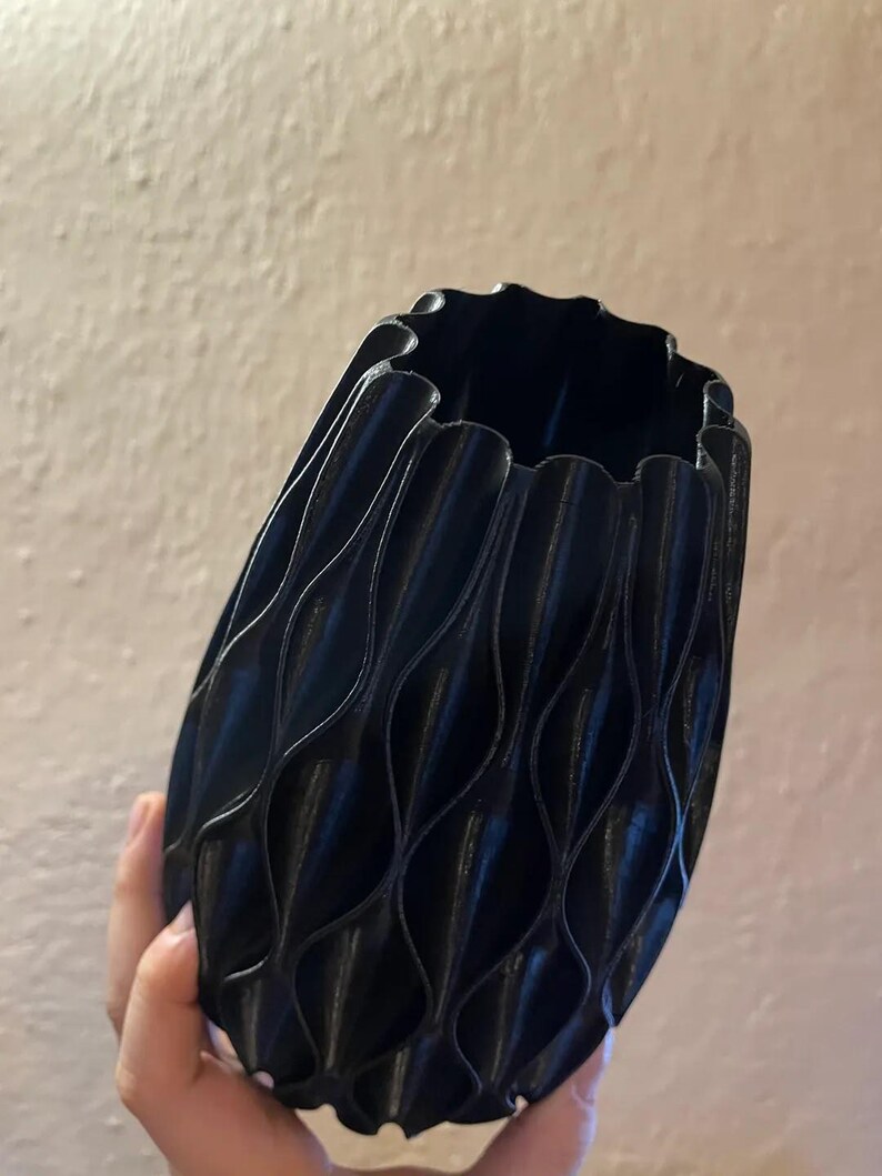 May include: A black vase with a repeating vertical wave pattern along its exterior surface. The vase features a scalloped rim and a rounded, bulbous body shape. The texture of the object appears to have fine horizontal lines consistent with 3D printing.
