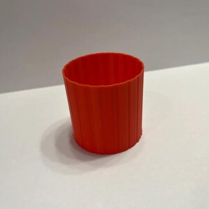 Op de afbeelding: Een feloranje, cilindervormige beker met verticale ribbels. De beker is gemaakt van een stevig materiaal en staat tegen een witte en grijze achtergrond. Het ontwerp van de beker suggereert dat het een herbruikbare koffiebekerhoes kan zijn.