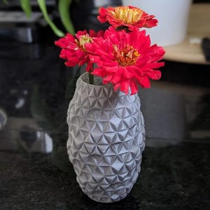 Op de afbeelding: Een grijze vaas met een geometrisch patroon bevat felrode en oranje bloemen. De vaas heeft een gestructureerd oppervlak met een herhalend driehoekig ontwerp. De bloemen hebben felrode bloemblaadjes en gele centra.