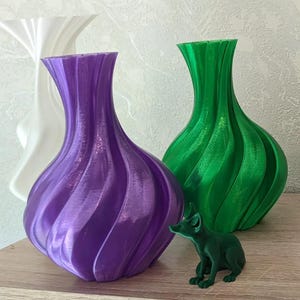 Op de afbeelding: Drie decoratieve vazen in wit, paars en groen, met een klein groen vossenbeeldje. De vazen hebben een uniek, geribbeld ontwerp. De paarse vaas staat op de voorgrond, met de groene vaas erachter. Het vossenbeeldje staat voor de groene vaas.