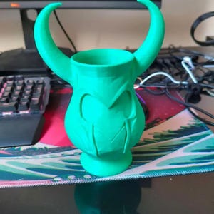 May include: A bright green decorative vase with curved horns and a stylized face design. The vase has a wide opening and a pedestal base. The horns curve upwards and outwards from the top of the vase. The design is reminiscent of a character from a comic book.