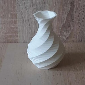 Op de afbeelding: Een witte, decoratieve vaas met een uniek spiraalontwerp. De vaas heeft een brede basis die taps toeloopt naar een smalle hals en een facetrand. Het is gemaakt van een solide, ondoorzichtig materiaal en is ongeveer 15 cm hoog.