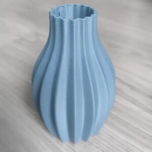 Op de afbeelding: Een lichtblauwe vaas met een getextureerd, geribbeld ontwerp. De vaas heeft een brede, afgeronde basis die naar boven toe smaller wordt, met een gegolfde rand. De vaas is gemaakt van een stevig materiaal en is geschikt voor het tentoonstellen van bloemen of als decoratief item.