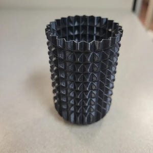 Può includere: Un contenitore cilindrico nero con un motivo geometrico strutturato. Il contenitore presenta sporgenze a forma di diamante che ricoprono la superficie. Il bordo superiore ha un design smerlato. L'oggetto è probabilmente stampato in 3D.