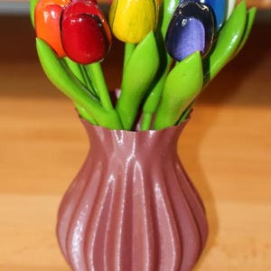 Könnte beinhalten: Eine dekorative Vase gefüllt mit bunten Holz-Tulpen. Die Tulpen sind in verschiedenen Farben bemalt, darunter Rot, Orange, Gelb, Rosa, Blau und Lila. Die Vase hat eine gedämpfte mauve Farbe und ein geripptes Design.