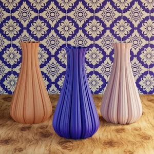 May include: Three decorative vases in peach, blue, and pink. The vases have a ribbed texture and a flared top. They are arranged on a wooden surface against a patterned blue and white background.