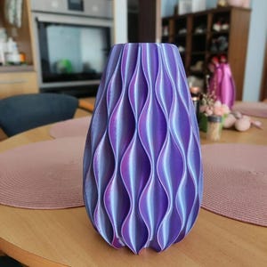May include: A vase with a vertical wave pattern featuring a gradient of iridescent purple and blue colours. The object has a cylindrical shape with a tapered top and a textured surface created by repeating concave and convex vertical ridges.