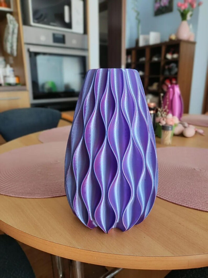 May include: A vase with a vertical wave pattern featuring a gradient of iridescent purple and blue colours. The object has a cylindrical shape with a tapered top and a textured surface created by repeating concave and convex vertical ridges.