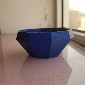 Puede incluir: Una maceta azul geométrica y facetada. La maceta tiene una abertura ancha y una superficie texturizada. La maceta está diseñada con bordes rectos y ángulos, creando una estética moderna. Es adecuada para plantas pequeñas o fines decorativos.