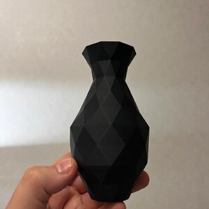 Op de afbeelding: Een zwarte geometrische vaas met een facetontwerp. De vaas heeft een brede basis die taps toeloopt naar een smallere hals en een uitlopende bovenkant. De vaas wordt in een hand gehouden.