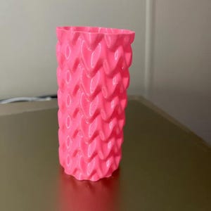 Op de afbeelding: Een felroze, cilindervormige vaas met een textuur, hartvormig patroon. De vaas is gemaakt van een stevig materiaal en staat rechtop op een reflecterend oppervlak. Het ontwerp heeft herhaalde, afgeronde vormen.