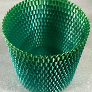 May include: A green, cylindrical container with a textured, geometric pattern. The container has a flared top and a solid base. The surface is covered in a repeating, scale-like design, creating a visually interesting texture. The colour is a vibrant, medium green.