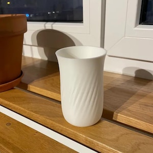 Op de afbeelding: Een witte keramische beker met een textuur, wervelend ontwerp. De beker is cilindrisch met een licht uitlopende bovenkant. Hij staat op een houten oppervlak bij een raam, met een terracotta pot op de achtergrond.