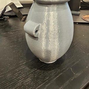 Puede incluir: Un jarrón gris impreso en 3D con un cuerpo redondeado y un borde ancho ligeramente ensanchado. Cuenta con un pequeño asa cilíndrica en el lateral. El jarrón tiene una superficie texturizada y un interior azul claro.