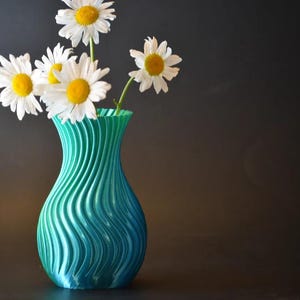 Op de afbeelding: Een turquoise en blauwe vaas met een golvend ontwerp bevat verschillende witte madeliefjes met gele centra. De vaas is het middelpunt, tegen een donkere achtergrond, waardoor een contrast ontstaat dat de bloemen benadrukt.