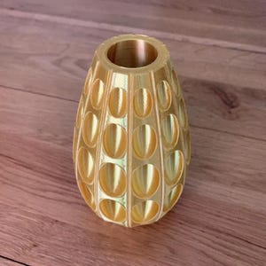 Op de afbeelding: Een goudkleurige, decoratieve vaas met een uniek, gestructureerd ontwerp. De vaas heeft een ovale vorm met een reeks ovale uitsparingen rond de buitenkant. De bovenste opening is cirkelvormig. De vaas is gemaakt van een glanzend, metalen materiaal.