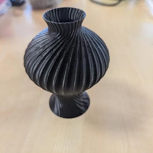 Op de afbeelding: Een zwarte decoratieve vaas met een textuur, geribbeld ontwerp. De vaas heeft een bolvormig lichaam en een uitlopende bovenkant, rustend op een voetstuk. De vaas is gemaakt van een solide, donker materiaal.