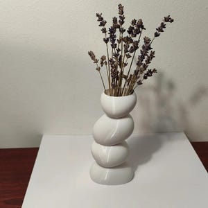 Può includere: Un vaso bianco a forma di pietre impilate che contiene lavanda essiccata. Il vaso ha una finitura liscia e lucida e un design moderno unico. La lavanda aggiunge un elemento naturale e terroso all'arredamento. Il vaso è alto circa 25 cm.
