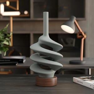 Puede incluir: Un jarrón moderno y abstracto en color gris claro, con un diseño en espiral. El jarrón se apoya sobre una base redonda de madera marrón. El fondo incluye una lámpara de escritorio y una estantería, sugiriendo un tema de decoración del hogar.