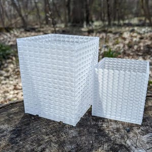 May include: Two clear, square planters with a woven texture sit on a weathered wooden surface. The larger planter is taller than the smaller one. The background features a blurred view of a forest.