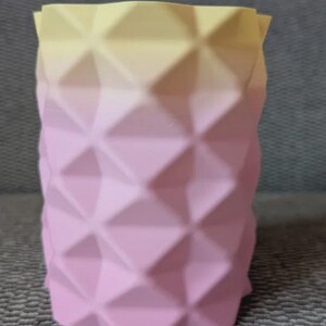 Op de afbeelding: Een decoratieve, geometrische vaas met een ruitpatroon. De vaas loopt van lichtgeel aan de bovenkant over in zachtroze aan de onderkant. De vaas is ongeveer 13 cm hoog.