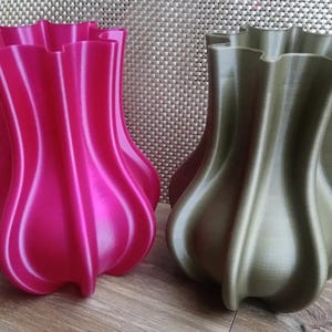 Op de afbeelding: Twee decoratieve vazen in tulpenvorm, één in fel magenta, de andere in gedempt olijfgroen. Beide hebben een glad, glanzend oppervlak met een gerimpelde rand. Geplaatst op een houten oppervlak.