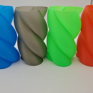 Op de afbeelding: Vier kleurrijke, gedraaide vazen in blauw, grijs, groen en oranje. De vazen hebben een uniek, spiraalvormig ontwerp en zijn gemaakt van een glad materiaal. Deze decoratieve vazen zijn geschikt voor woondecoratie.