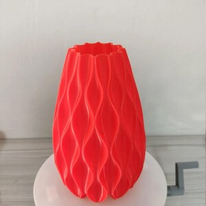 May include: A bright red vase with a repeating vertical wave pattern along its exterior. The vase has a scalloped top edge and a tapered cylindrical shape, featuring a consistent matte finish across its textured, undulating surface.