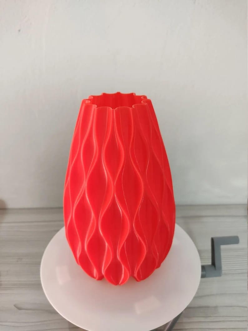 May include: A bright red vase with a repeating vertical wave pattern along its exterior. The vase has a scalloped top edge and a tapered cylindrical shape, featuring a consistent matte finish across its textured, undulating surface.
