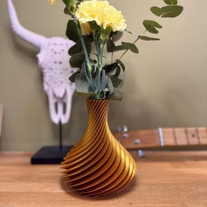 Op de afbeelding: Een goudkleurige, spiraalvormig ontworpen vaas bevat gele anjers en groene eucalyptusbladeren. De vaas staat op een houten oppervlak. Op de achtergrond staat een witte decoratieve stierschedel.
