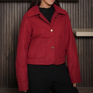 May include: A red jacket with a classic collar, two front pockets, and gold buttons. The jacket is a solid color and appears to be made of a wool or wool-blend fabric. The jacket is paired with black pants.