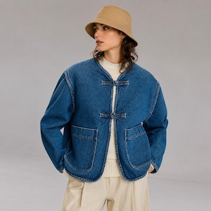 May include: A tan bucket hat worn with a blue denim jacket featuring a collarless neckline, two front patch pockets, and two toggle-style closures down the center front. The jacket is layered over a cream-colored ribbed knit sweater and light trousers.