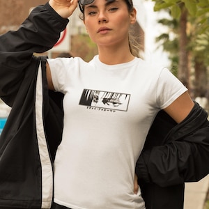 May include: White t-shirt featuring a black and white anime eye graphic and Japanese text. The person is wearing black sunglasses and a black jacket. The background includes palm trees and a pavement.