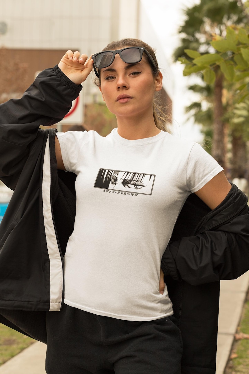 May include: White t-shirt featuring a black and white anime eye graphic and Japanese text. The person is wearing black sunglasses and a black jacket. The background includes palm trees and a pavement.