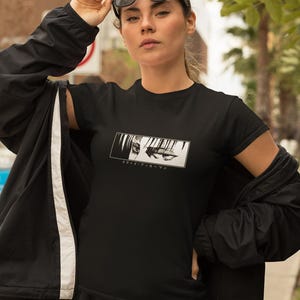 May include: Black t-shirt with a rectangular graphic of anime eyes and Japanese text. The short-sleeved shirt is worn with a black jacket and trousers. The person is wearing black sunglasses.