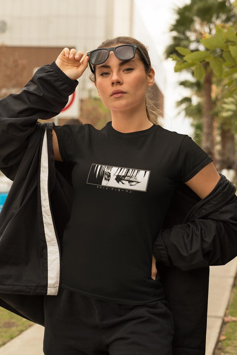May include: Black t-shirt with a rectangular graphic of anime eyes and Japanese text. The short-sleeved shirt is worn with a black jacket and trousers. The person is wearing black sunglasses.