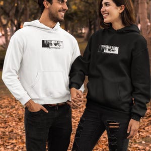 May include: A white and a black hoodie, each featuring a black and white graphic of eyes and text. Both hoodies have a hood and a front pocket. The image shows two people wearing the hoodies.