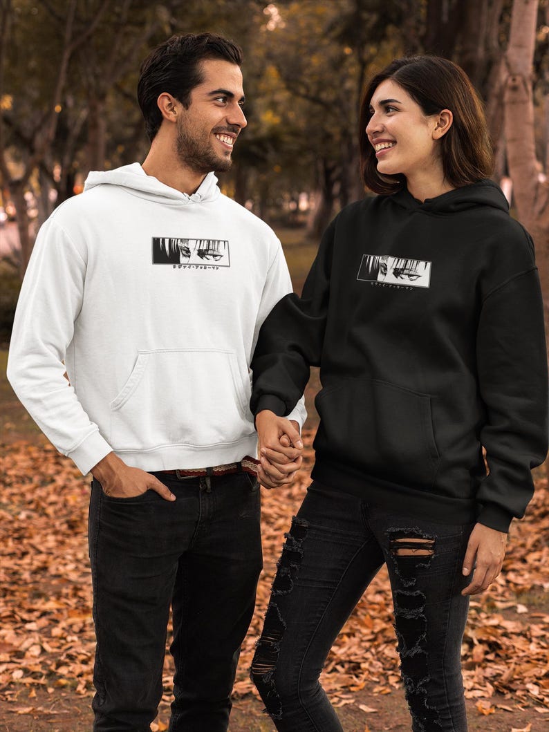 May include: A white and a black hoodie, each featuring a black and white graphic of eyes and text. Both hoodies have a hood and a front pocket. The image shows two people wearing the hoodies.