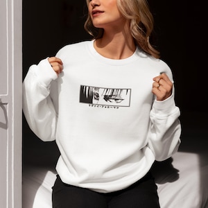 May include: White crew neck sweatshirt with a black and white anime eye graphic in a rectangular frame. Japanese text is below the graphic. The sweatshirt is made of a soft material.