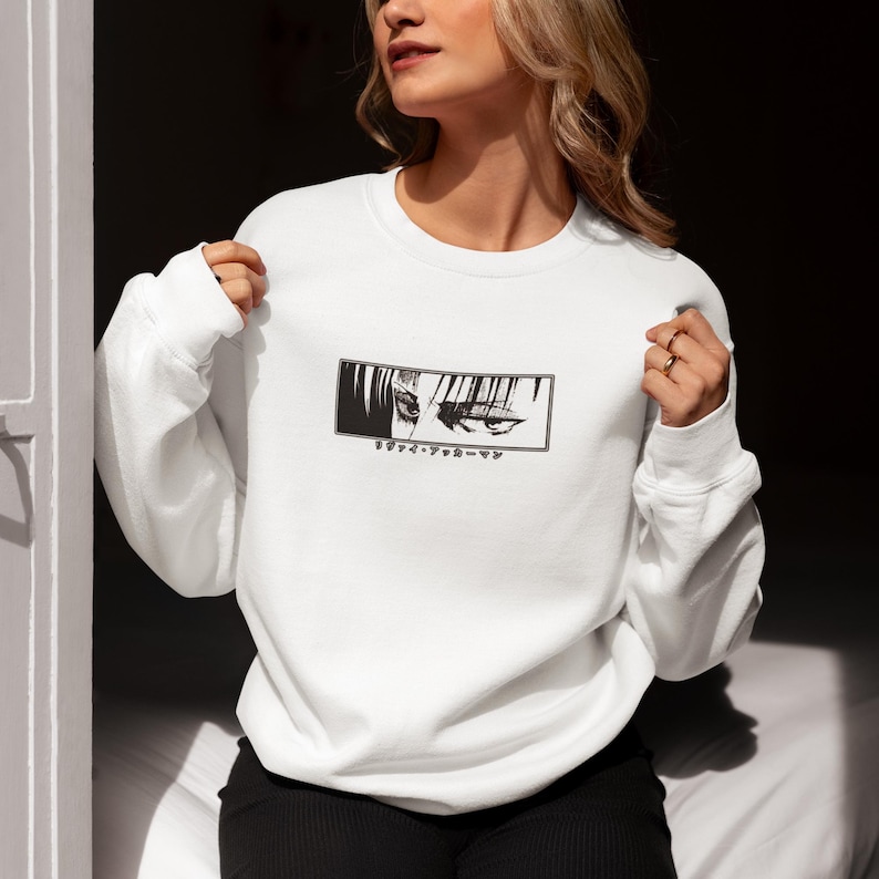 May include: White crew neck sweatshirt with a black and white anime eye graphic in a rectangular frame. Japanese text is below the graphic. The sweatshirt is made of a soft material.