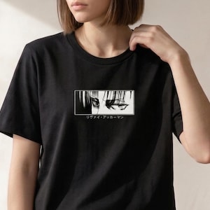 May include: Black t-shirt featuring a rectangular graphic of anime eyes in black and white. Below the graphic is Japanese text. The shirt is a crew neck and short-sleeved.
