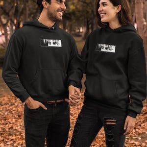 May include: Two people wearing black hoodies with a white graphic design. The hoodies have a front pocket and a hood. They are holding hands and smiling. They are wearing black jeans. The background is a blurred outdoor scene with trees and fallen leaves.