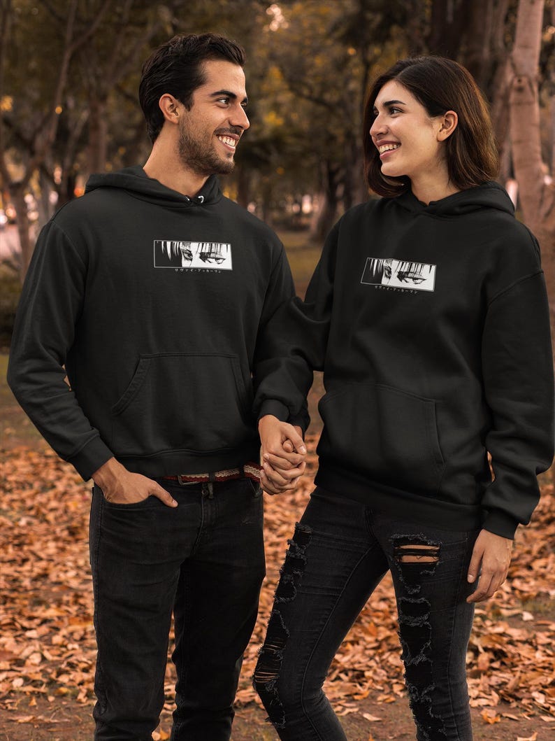 May include: Two people wearing black hoodies with a white graphic design. The hoodies have a front pocket and a hood. They are holding hands and smiling. They are wearing black jeans. The background is a blurred outdoor scene with trees and fallen leaves.